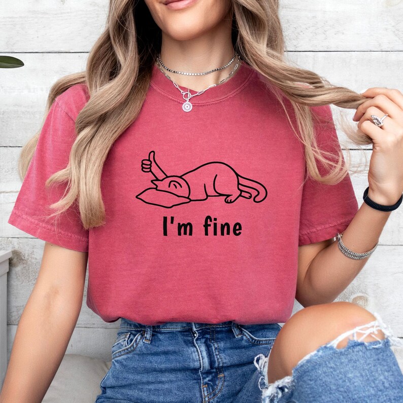 May include: A maroon t-shirt featuring the text "I'm fine" beneath a black line drawing of a cat giving a thumbs up. The cat is lying on a pillow. The shirt is a plain colour.
