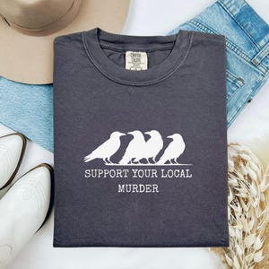 Puede incluir: Camiseta gris oscuro con un gr&aacute;fico blanco de cuatro cuervos y el texto "SUPPORT YOUR LOCAL MURDER". La camiseta est&aacute; doblada y se muestra con un sombrero, vaqueros y botas blancas.