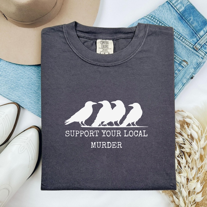 Puede incluir: Camiseta gris oscuro con un gr&aacute;fico blanco de cuatro cuervos y el texto "SUPPORT YOUR LOCAL MURDER". La camiseta est&aacute; doblada y se muestra con un sombrero, vaqueros y botas blancas.