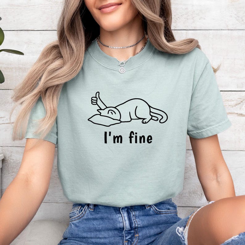 May include: A pale green t-shirt featuring a black cartoon cat giving a thumbs up, with the words "I'm fine" underneath. The shirt has a crew neck and short sleeves.