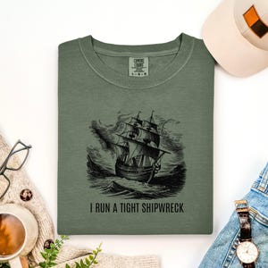 Könnte beinhalten: Olivgrünes Sweatshirt mit einem schwarzen Bild eines Schiffes auf stürmischer See. Darunter steht der Text "I RUN A TIGHT SHIPWRECK". Das Sweatshirt ist von der Marke Comfort Colors.