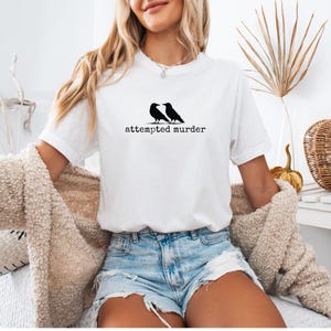 Pu&ograve; includere: T-shirt bianca con grafica nera di due corvi e la scritta "attempted murder" in un font semplice. La maglietta &egrave; abbinata a pantaloncini di jeans e un cardigan beige.