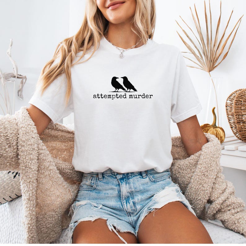 Pu&ograve; includere: T-shirt bianca con grafica nera di due corvi e la scritta "attempted murder" in un font semplice. La maglietta &egrave; abbinata a pantaloncini di jeans e un cardigan beige.