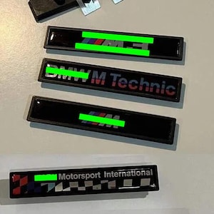 Pode incluir: Vários emblemas de carro pretos com texto e logotipos, incluindo "BMW M Technic" e "Motorsport International". Os emblemas têm um acabamento brilhante e vários detalhes coloridos, como vermelho, azul e branco. Uma braçadeira de montagem preta está no topo.