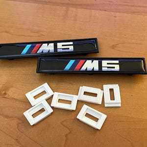 Puede incluir: Dos emblemas rectangulares negros para coche con el texto "M5" en blanco y un diseño de rayas azules, rojas y celeste. También se ven seis clips rectangulares blancos.
