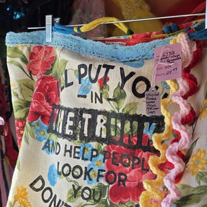 May include: A floral print tote bag with the text "I'll put you in the trunk and help people look for you, don't test me." The bag has a blue crocheted trim and colorful crocheted accents.