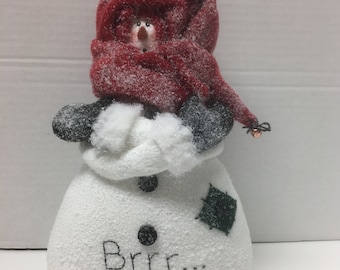 May include: A white snowman figurine with a red hat and scarf, black eyes, a carrot nose, and a black button. The snowman is holding its arms in front of it and has the text "Brrr..." written on its front.