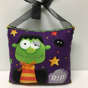 May include: A Halloween-themed decorative pillow featuring a green Frankenstein's monster design on a purple background. The pillow has a black ribbon for hanging, a spider, stars, and a tombstone with "RIP" on it. The pillow is approximately 10 inches square.