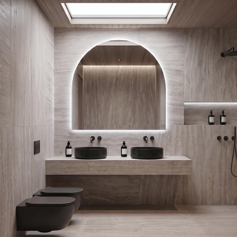 May include: A modern bathroom with a neutral color palette. The focal point is a large, arched mirror with integrated lighting. Two black vessel sinks sit atop a stone countertop. The room includes a toilet, shower, and wooden ceiling.