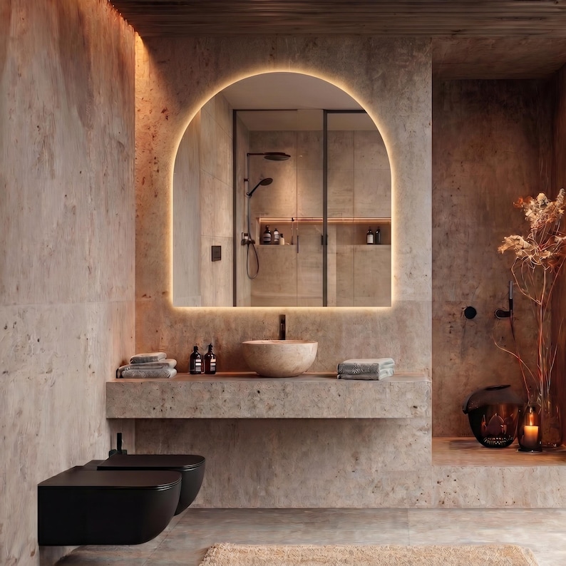 May include: A modern bathroom with a large, arched mirror with a warm, glowing light. The room features a stone countertop with a vessel sink, towels, and toiletries. A black toilet and bidet are in the foreground. The color palette is neutral with warm tones.