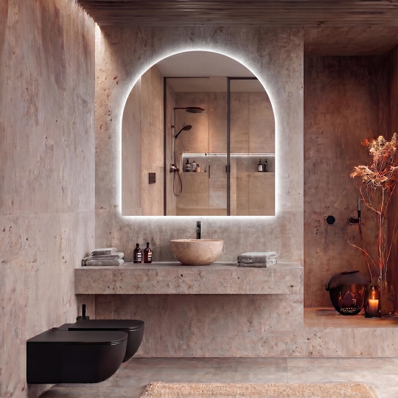 May include: A modern bathroom with a large, arched mirror with a glowing border. The room features a stone sink, a concrete shelf with towels and bottles, and a black toilet. The color palette is neutral with warm tones.
