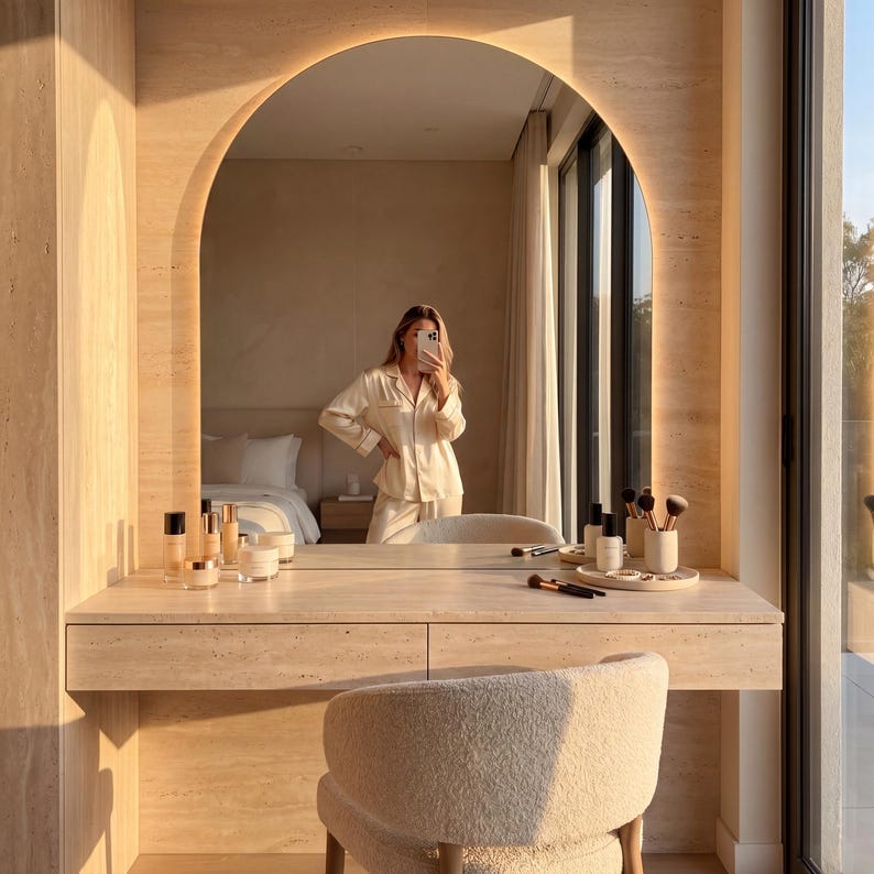 May include: A beige vanity with a large arched mirror and built-in lighting. The vanity has a drawer and a cream-colored chair. Makeup brushes and bottles are arranged on the vanity. A person in a cream-colored pajama set is reflected in the mirror.