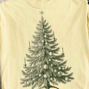 May include: A pale yellow t-shirt featuring a centred, detailed dark green illustration of a Christmas tree. The tree is decorated with round ornaments and several upright candles, topped with a star. The design has a vintage, etched aesthetic.