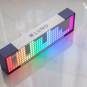 Puede incluir: Una pantalla LED rectangular con la palabra "LUNO" y un patrón de ecualizador gráfico. La pantalla presenta un cuerpo blanco con tapas de color azul oscuro y una vibrante gama de colores, incluyendo rojo, naranja, verde y azul.