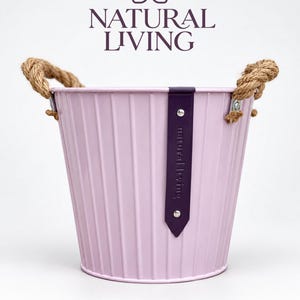 May include: A light purple metal bucket with vertical ridges, featuring two thick twisted jute rope handles attached with silver rivets. A dark purple vertical leather-like strap is centred on the front, embossed with the text Natural Living.