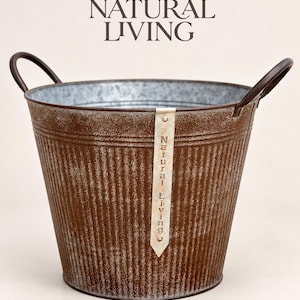 May include: A brown, corrugated metal bucket with two dark handles and a vertical metal tag featuring the text Natural Living. The bucket has a wide, open top and a tapered base, with a weathered, textured finish throughout the exterior surface.