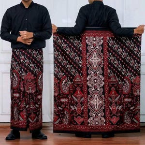 以下が含まれることがあります： 赤、黒、白の色合いの模様入りサロン。生地は、花柄と幾何学模様の複雑なデザインが特徴です。サロンは、黒いシャツとズボンを着ている人に表示されています。