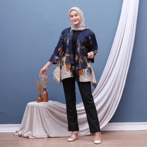 Op de afbeelding: Een vrouw draagt een marineblauwe en beige jas met bloemenprint, een zwarte broek en een beige hoofdbedekking. De jas heeft een asymmetrische zoom. De outfit wordt gecombineerd met beige hakken. De achtergrond is een effen blauwe muur.