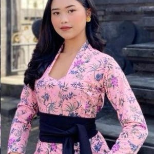 May include: A pink floral top with long sleeves and a V-neck, paired with a black sash and skirt. The top features a pattern of purple and green leaves and flowers. A gold ring and earrings complete the look.