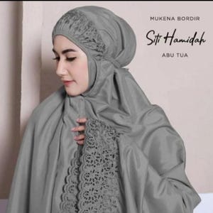 May include: A grey prayer garment, or mukena, with detailed lace embellishments. The head covering and sleeves are decorated with delicate lace patterns. The image includes the text "MUKENA BORDIR", "Siti Hamidah", and "ABU TUA".