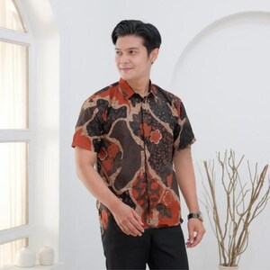 Puede incluir: Camisa de manga corta con estampado batik en tonos marrón, negro y rojo. La camisa presenta un diseño floral y abstracto. El modelo lleva pantalones negros y un reloj.