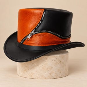 Puede incluir: Un sombrero de copa construido con paneles de cuero negro y naranja. El sombrero presenta una prominente cremallera metálica de color plateado que se curva diagonalmente por el lateral, separando las secciones naranja y negra. Tiene un ala ancha y curva.