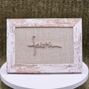 May include: A framed artwork with the word "faith" spelled out in silver-colored script, embellished with rhinestones. The artwork is set against a beige linen background and framed in a distressed white wood frame.
