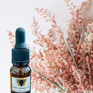 May include: An amber glass bottle of Bee Serum with a black dropper, beside a bunch of pink flowers. The label reads "Honey Springs Bee" and "Bee Venom Apitoxin". The text "Bee Serum" and "with Royal Jelly and Manuka Honey" are at the top.