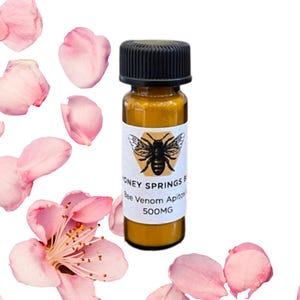 Könnte beinhalten: Kleine bernsteinfarbene Glasflasche mit schwarzem Deckel, beschriftet mit "HONEY SPRINGS B Bee Venom Apitox 500MG". Rosa Blütenblätter und Blüten umgeben die Flasche. Der Text "Bee Powder" befindet sich oben im Bild.