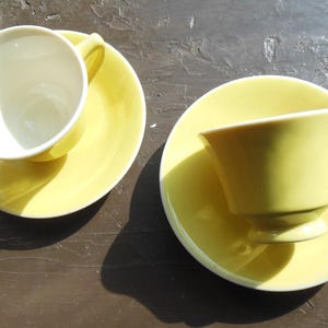 May include: Two yellow teacups and saucers are arranged on a dark surface. One cup is upright, revealing a white interior, while the other is inverted. The saucers and cups are a pale yellow color, creating a simple, elegant aesthetic.