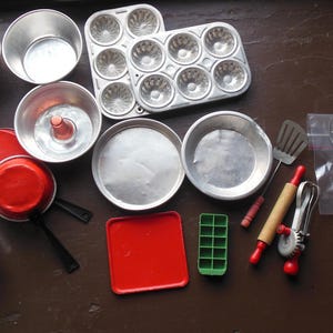 May include: A collection of miniature kitchenware including aluminum pots, pans, and a muffin tin. The set features red and black plastic-handled utensils, a small rolling pin, a green ice cube tray, a red square tray, and a plastic bag of silverware.