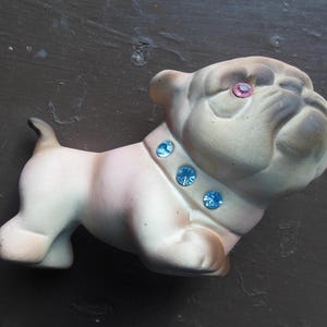 May include: A ceramic bulldog figurine in shades of cream and pink, with a pink gem eye and a collar adorned with three blue gems. The bulldog is posed in a standing position.