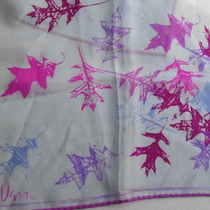 May include: A square, light blue scarf with a purple border and leaf pattern. The leaves are in shades of pink, purple, and blue. The scarf has the word "Usna" in the bottom left corner.