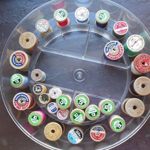 May include: A circular, clear plastic storage tray containing numerous vintage sewing thread spools. The spools are made of wood and plastic, featuring various colors including red, blue, green, and white, with labels from brands like Coats & Clark.