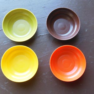 May include: A set of four small, round, smooth bowls in solid colors. The collection includes one lime green bowl, one dark brown bowl, one bright yellow bowl, and one vibrant orange bowl. Each bowl features a simple, circular design.