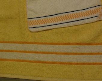 May include: A yellow bath towel with white and orange stripes at the bottom. A smaller, off-white hand towel with orange and black geometric patterns is placed on top. The towels are made of a soft, absorbent material.
