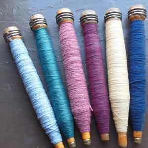 May include: A collection of six vintage wooden spools, each wrapped with colorful yarn. The spools are arranged diagonally, showcasing shades of blue, teal, pink, purple, cream, and navy. The spools have a wooden base and a metal top.