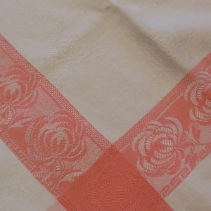 May include: Cream-colored tablecloth with a coral-colored floral lace border. The border features a repeating rose pattern. The tablecloth is folded, creating a V-shape design.