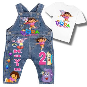 Pode incluir: Macacão de ganga e uma camiseta branca com personagens da Dora, a Aventureira. O macacão tem o nome "KAYLA" e o número "2" em rosa, juntamente com gráficos de personagens. A camiseta mostra Dora e Botas.