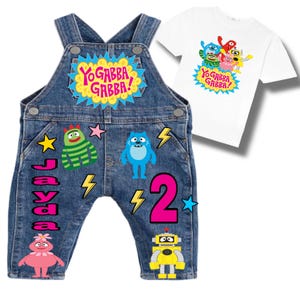 May include: Denim overalls with colorful character patches and the text "Jayda" and a large pink "2". A white t-shirt with a "Yo Gabba Gabba!" graphic is next to the overalls. The overalls have adjustable straps.