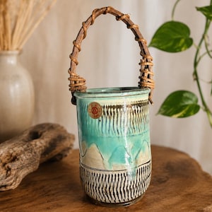 May include: A ceramic vase with a woven handle. The vase features a turquoise and cream color scheme with a black and white patterned base. The handle is made of woven brown material.