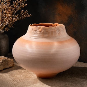 May include: A handcrafted, light-colored ceramic vase with a wide, rounded body and a textured, wavy rim. The vase features a band of orange-pink color around the middle. The vase is a decorative item.