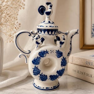 May include: White and blue ceramic teapot with a rooster-shaped lid. The teapot features a circular design with blue floral patterns and a curved handle. The spout is elegantly curved, and the base is pedestal-style. The teapot is a decorative item.