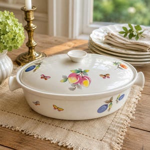 Könnte beinhalten: Eine ovale Servierschüssel aus weißer Keramik mit Deckel, verziert mit Illustrationen von Pfirsichen, Pflaumen und Schmetterlingen. Die Schüssel hat zwei Griffe und einen kleinen Knopf auf dem Deckel. Sie steht auf einem beigen Platzset.