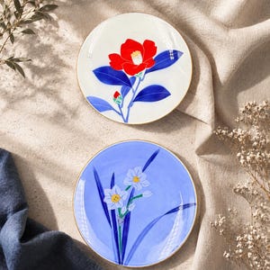 May include: Two decorative ceramic plates with floral designs. One plate features a red flower with blue leaves on a white background. The other plate has white flowers with yellow centres and blue leaves on a light blue background.
