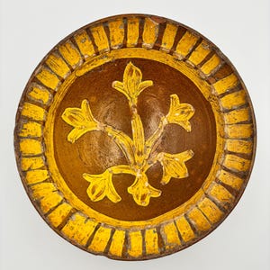 Puede incluir: Un plato decorativo redondo de cerámica con un centro marrón y un borde amarillo. El centro presenta un diseño floral amarillo. El borde tiene un patrón rectangular amarillo repetido. El plato tiene un aspecto rústico y envejecido.