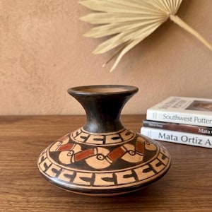 May include: A handcrafted, ceramic vase with a dark brown neck and a beige body. The body features a repeating geometric pattern in black and reddish-brown. The vase is displayed on a wooden surface, with books in the background.