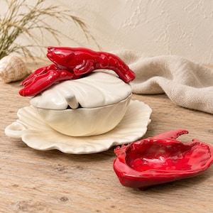 Puede incluir: Un cuenco de cerámica blanca con tapa y plato a juego, que presenta un asa de langosta roja esculpida en la tapa. Una pequeña cuchara blanca descansa dentro del cuenco. Junto al cuenco hay un plato de cerámica roja con forma de pinza de langosta.