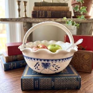 Könnte beinhalten: Ein weißer Keramikkorb mit Henkel, gewelltem Rand und blauem Blumenmuster. Der Korb enthält einen roten Apfel, einen grünen Apfel und einen Silberlöffel. Er steht auf einem Stapel blauer Bücher.