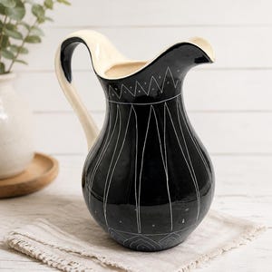 May include: A ceramic pitcher with a glossy black exterior and a cream-coloured interior and handle. The black surface features hand-etched white line patterns, including vertical stripes, a zigzag border near the rim, and geometric shapes at the base.